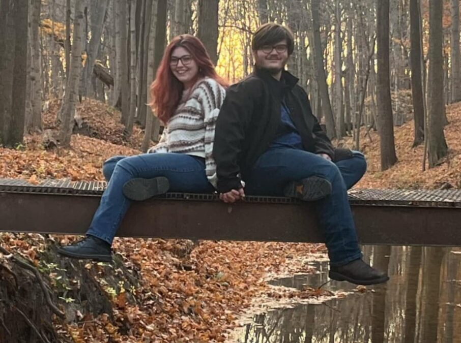 Elise and Emily Two young women sitting together on a large wooden log or beam outdoors in a wooded area with autumn leaves on the ground.